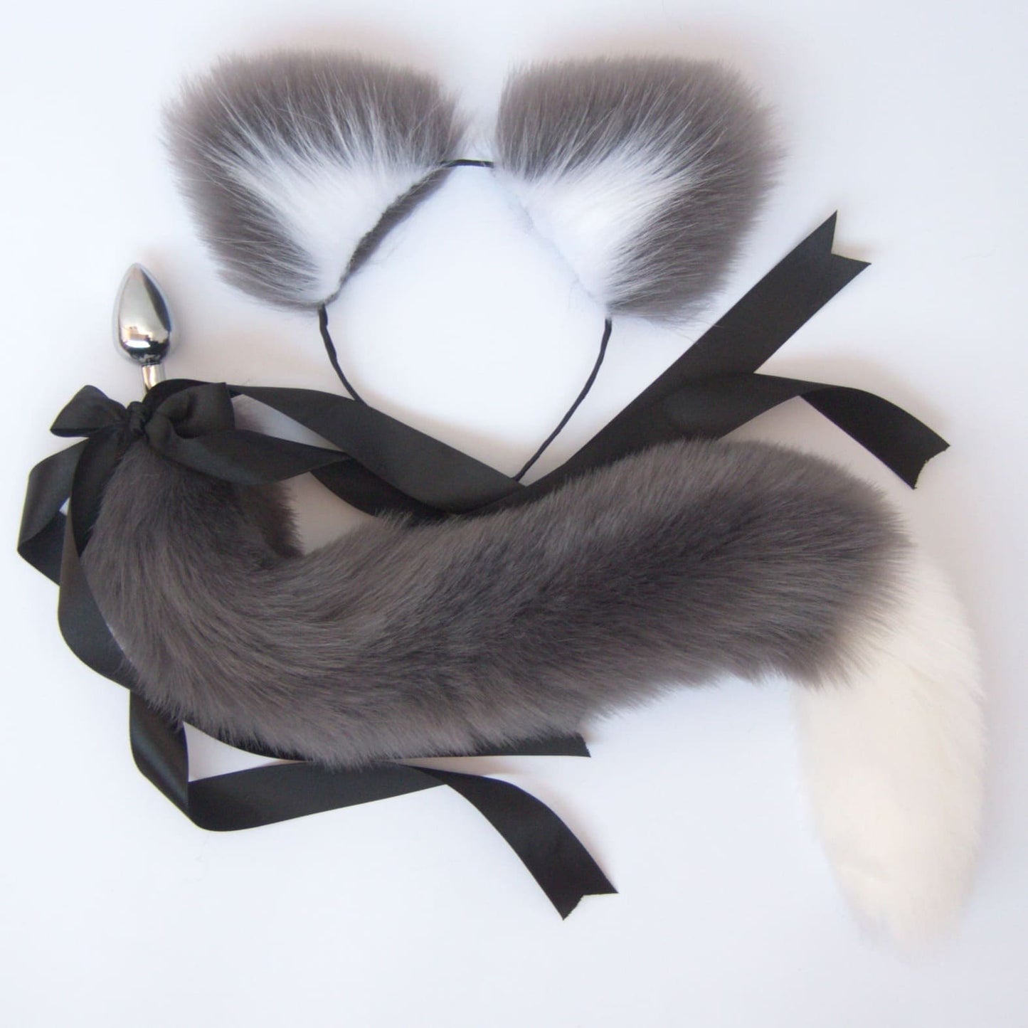 A gray and white faux fur cat tail butt plug with a stainless steel plug and a black ribbon attached.